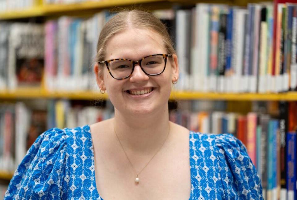 Katherine Fallon-Reusch smiling, wearing glasses, and standing in front of a library bookshelf